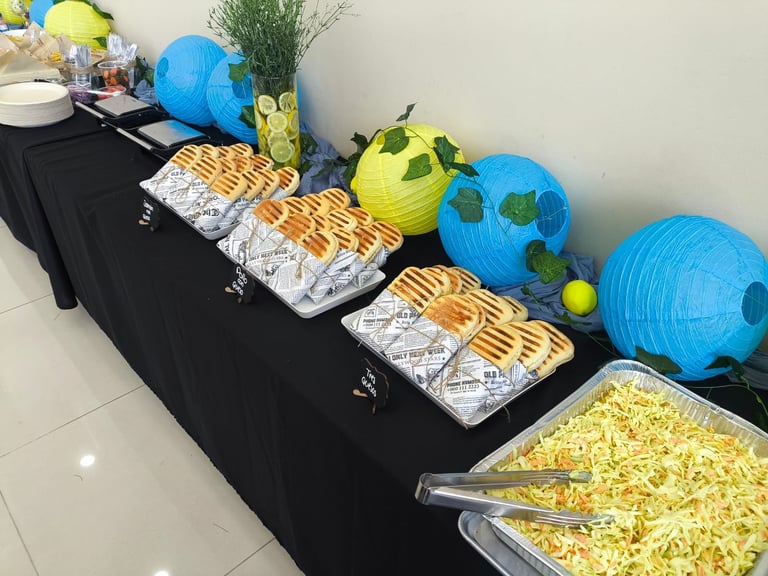 Party snack table display with blue and yellow decorative balls, foil trays of food, popcorn, and green decorative items on black tablecloth