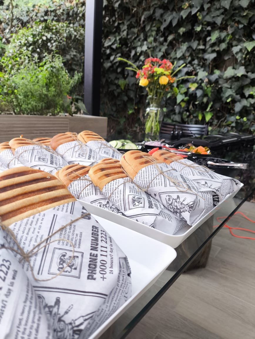 Bakery items including bagels and pastries wrapped in newspaper on a table with fresh flowers in a vase outdoors