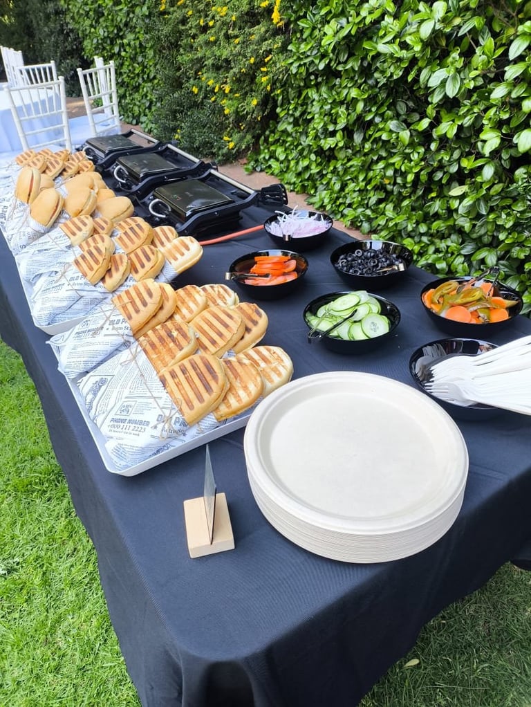 Outdoor buffet table with waffles, fresh vegetables, empty white plates, and various toppings on a black tablecloth